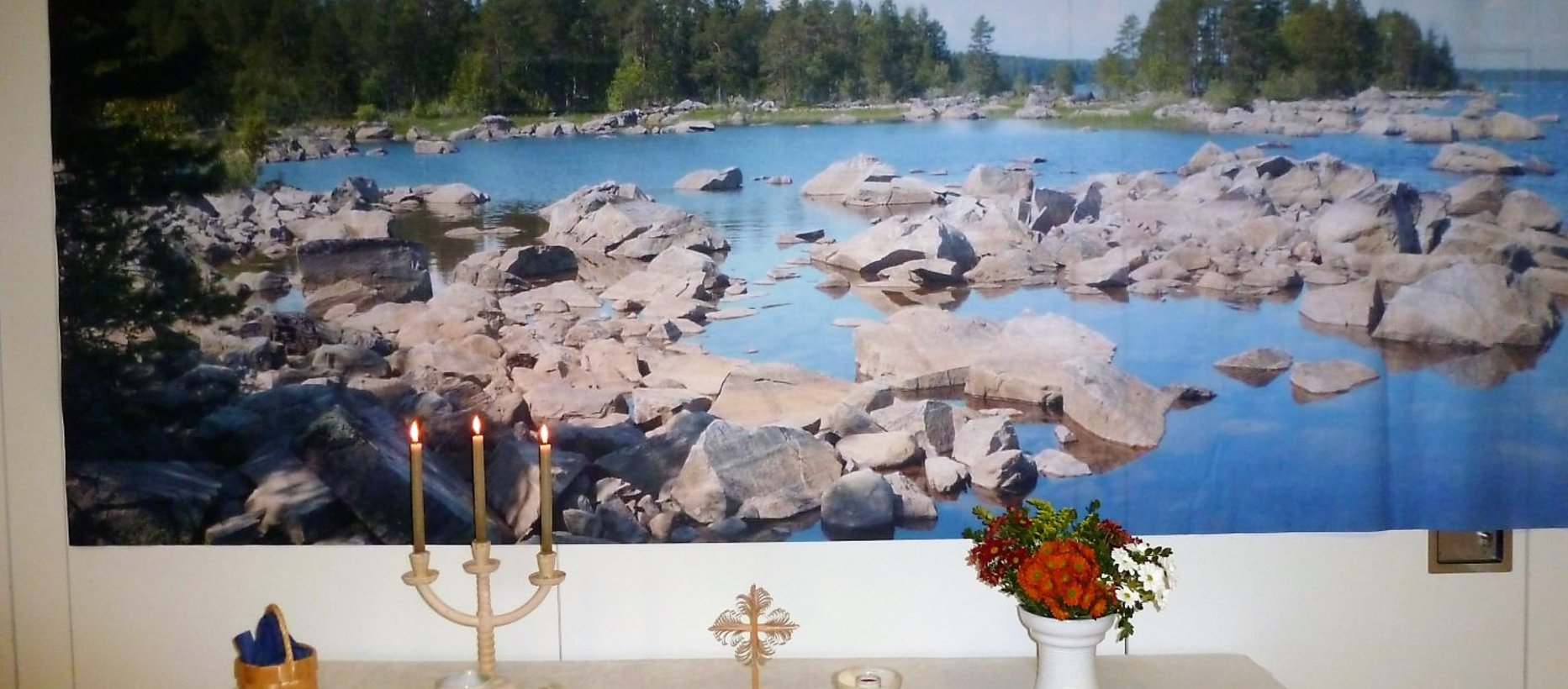 Bild mit einer steinigen Uferlandschaft, Kerzenständer mit drei Kerzen, Holzkreuz, Blumen in einer Vase