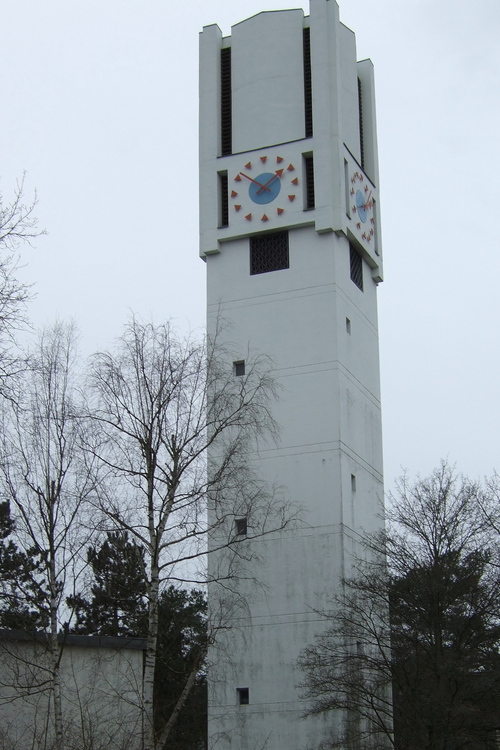 Der moderne Kirchturm ist weiß, aus Beton und hat eine Uhr.
