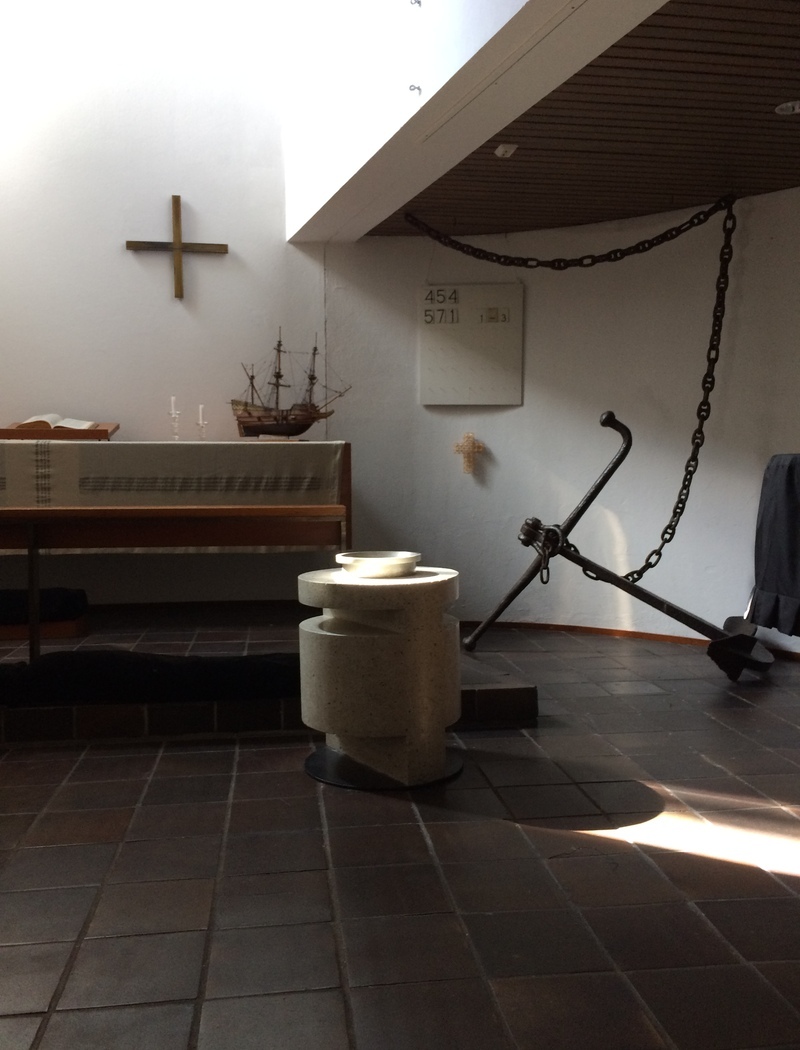 Im Kirchenraum im Vordergrund ein moderner Taufstein, dahinter ein großer Anker.